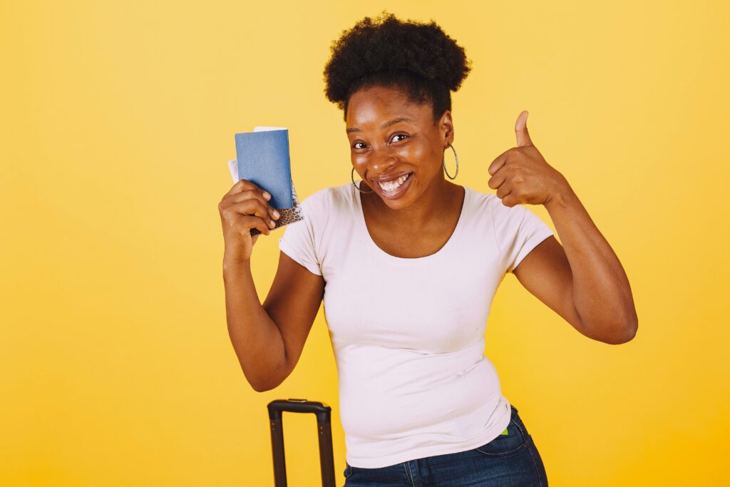Smiling woman holding passport giving thumbs up against yellow background.
